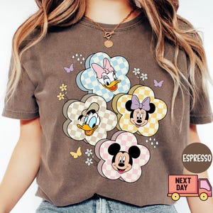 May include: Espresso-colored t-shirt featuring a floral design with Disney characters Daisy Duck, Donald Duck, Minnie Mouse, and Mickey Mouse. The flowers have checkered patterns in pastel colors. Butterflies and small flowers are also included in the design.