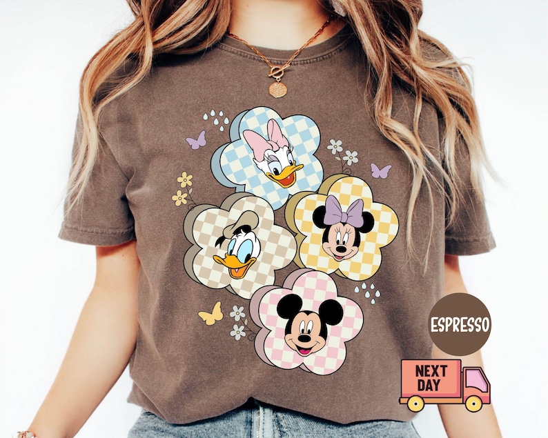 May include: Espresso-colored t-shirt featuring a floral design with Disney characters Daisy Duck, Donald Duck, Minnie Mouse, and Mickey Mouse. The flowers have checkered patterns in pastel colors. Butterflies and small flowers are also included in the design.