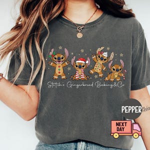 Puede incluir: Camiseta gris oscuro con un dise&ntilde;o de cinco personajes de dibujos animados con forma de pan de jengibre, cada uno con accesorios navide&ntilde;os &uacute;nicos. El texto "Stitch's Gingerbread Baking & Co" est&aacute; impreso debajo. Un cami&oacute;n de reparto rosa est&aacute; en la esquina inferior derecha.