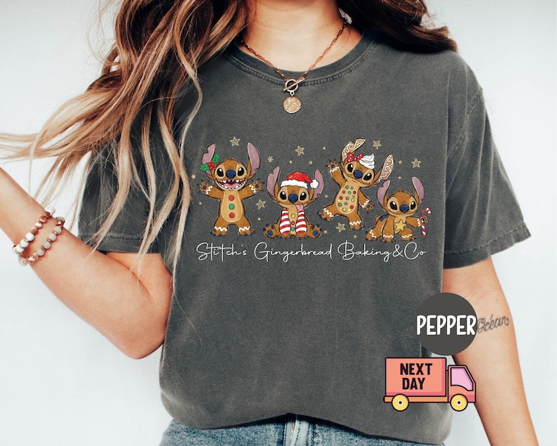 Puede incluir: Camiseta gris oscuro con un dise&ntilde;o de cinco personajes de dibujos animados con forma de pan de jengibre, cada uno con accesorios navide&ntilde;os &uacute;nicos. El texto "Stitch's Gingerbread Baking & Co" est&aacute; impreso debajo. Un cami&oacute;n de reparto rosa est&aacute; en la esquina inferior derecha.