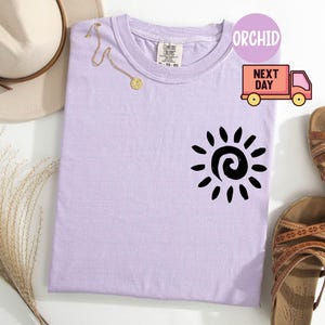 May include: A light purple t-shirt with a black sun graphic. The shirt is next to a pair of brown sandals, a gold necklace, and a beige hat. The shirt has the word "ORCHID" in a circle.