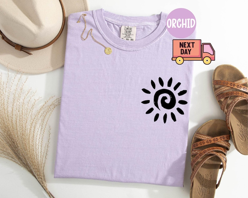 May include: A light purple t-shirt with a black sun graphic. The shirt is next to a pair of brown sandals, a gold necklace, and a beige hat. The shirt has the word "ORCHID" in a circle.
