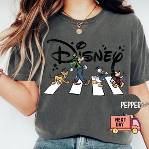 K&ouml;nnte beinhalten: Dunkelgraues T-Shirt mit dem Disney-Logo und den Zeichentrickfiguren Goofy, Donald Duck und Mickey Mouse, die eine Stra&szlig;e &uuml;berqueren. Das T-Shirt hat einen Vintage-Look.