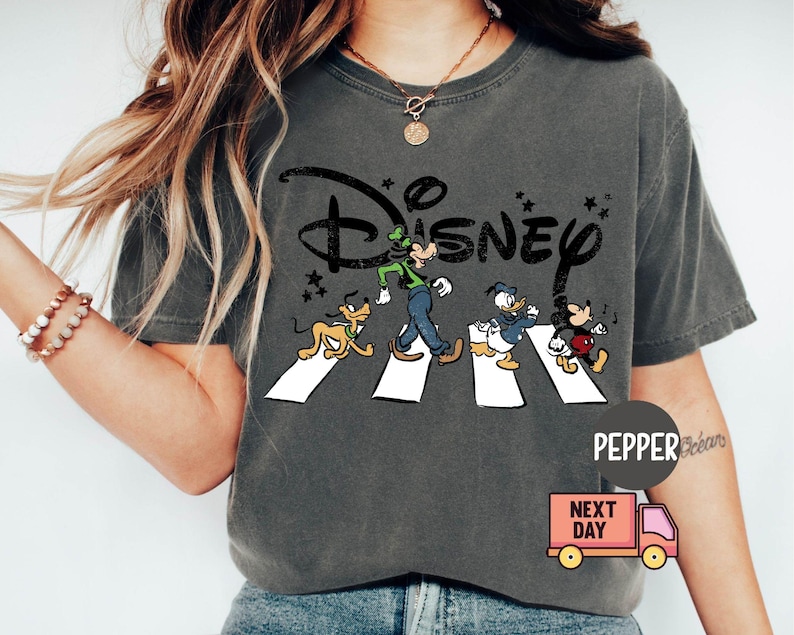 K&ouml;nnte beinhalten: Dunkelgraues T-Shirt mit dem Disney-Logo und den Zeichentrickfiguren Goofy, Donald Duck und Mickey Mouse, die eine Stra&szlig;e &uuml;berqueren. Das T-Shirt hat einen Vintage-Look.