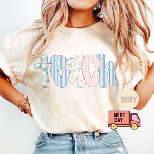 May include: Ivory-colored t-shirt with the word "TEACH" in a patchwork design. The letter "T" is a floral cross, "E" has bows, "A" is pink gingham, "C" and "H" are blue stripes. The shirt has rolled-up sleeves.