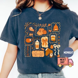 May include: Navy blue t-shirt with a colorful autumn-themed graphic. The design includes pumpkins, a slice of pie, a coffee cup, a candle, a bow, and other fall elements. The word "Autumn" is also featured.