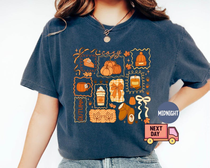 May include: Navy blue t-shirt with a colorful autumn-themed graphic. The design includes pumpkins, a slice of pie, a coffee cup, a candle, a bow, and other fall elements. The word "Autumn" is also featured.