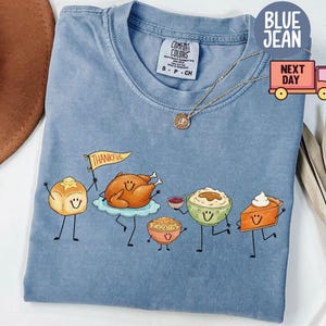 Puede incluir: Una camiseta de color azul vaquero con un gráfico de alimentos de Acción de Gracias antropomórficos, incluyendo un panecillo con una bandera que dice THANKFUL, un pavo asado, relleno, puré de patatas y una porción de tarta de calabaza.