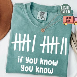 May include: Light green t-shirt with white text and tally marks. The phrase "if you know you know" is printed below the tally marks. The shirt has a crew neck and short sleeves. A gold necklace is draped over the shirt.