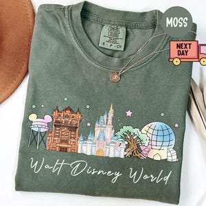 May include: Moss green t-shirt featuring a colorful graphic of Walt Disney World landmarks, including Cinderella's castle and the Hollywood Tower Hotel. The words "Walt Disney World" are written in a script font below the graphic.