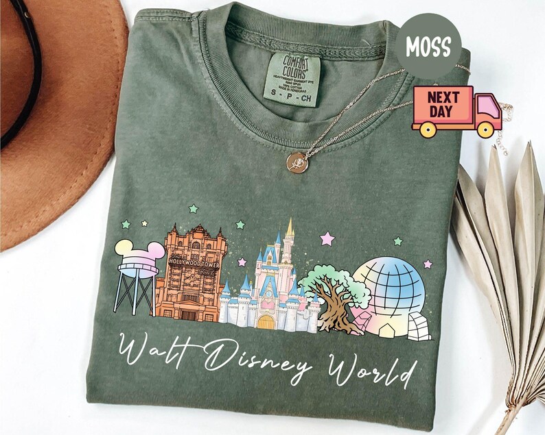 May include: Moss green t-shirt featuring a colorful graphic of Walt Disney World landmarks, including Cinderella's castle and the Hollywood Tower Hotel. The words "Walt Disney World" are written in a script font below the graphic.