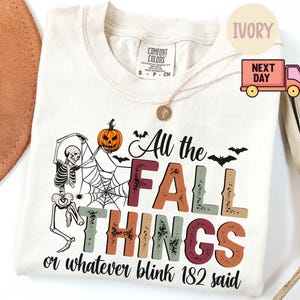 May include: Ivory-colored t-shirt with a graphic design featuring a skeleton, pumpkin, and spiderweb. The text reads "All the FALL THINGS or whatever blink 182 said" in various colors. A necklace and a hat are also visible.