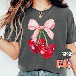 Puede incluir: Camiseta gris oscuro con una ilustraci&oacute;n de acuarela de tres cerezas rojas con un lazo rosa. La camiseta tiene un corte holgado y est&aacute; hecha de un material suave. El texto "PEPPER" es visible.