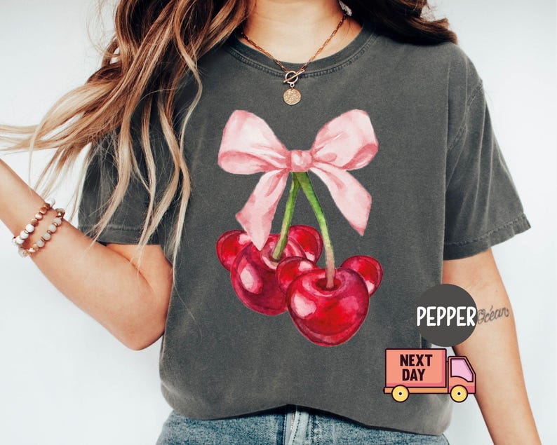 Puede incluir: Camiseta gris oscuro con una ilustraci&oacute;n de acuarela de tres cerezas rojas con un lazo rosa. La camiseta tiene un corte holgado y est&aacute; hecha de un material suave. El texto "PEPPER" es visible.