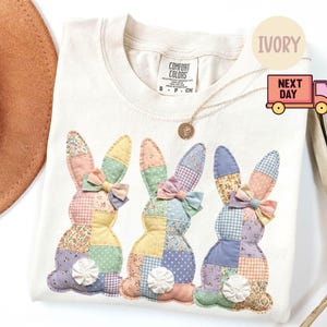 May include: Ivory-colored sweatshirt featuring three patchwork bunny designs with floral and gingham patterns. Each bunny has a bow and a decorative tail. The shirt has a crew neck and a Comfort Colors tag.