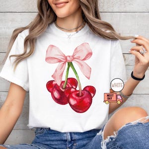 Puede incluir: Camiseta blanca con una ilustraci&oacute;n de acuarela de tres cerezas rojas y un lazo rosa. Las cerezas tienen tallos verdes. La camiseta tiene cuello redondo. El texto "WHITE" est&aacute; en un c&iacute;rculo blanco.