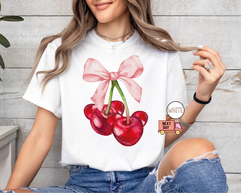 Puede incluir: Camiseta blanca con una ilustraci&oacute;n de acuarela de tres cerezas rojas y un lazo rosa. Las cerezas tienen tallos verdes. La camiseta tiene cuello redondo. El texto "WHITE" est&aacute; en un c&iacute;rculo blanco.