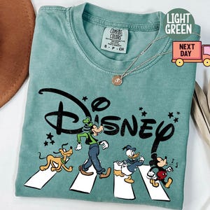 K&ouml;nnte beinhalten: Hellgr&uuml;nes T-Shirt mit dem Wort "Disney" in Schwarz und Zeichentrickfiguren, die einen Zebrastreifen &uuml;berqueren. Die Figuren sind Goofy, Pluto, Donald Duck und Mickey Mouse. Das Shirt hat einen Rundhalsausschnitt und kurze &Auml;rmel.