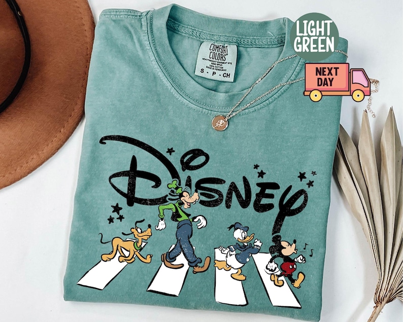 K&ouml;nnte beinhalten: Hellgr&uuml;nes T-Shirt mit dem Wort "Disney" in Schwarz und Zeichentrickfiguren, die einen Zebrastreifen &uuml;berqueren. Die Figuren sind Goofy, Pluto, Donald Duck und Mickey Mouse. Das Shirt hat einen Rundhalsausschnitt und kurze &Auml;rmel.