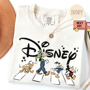 K&ouml;nnte beinhalten: Elfenbeinfarbenes T-Shirt mit dem Wort "Disney" in einer stilisierten Schriftart und Zeichentrickfiguren, die einen Zebrastreifen &uuml;berqueren. Zu den Figuren geh&ouml;ren Goofy, Pluto, Donald Duck und Mickey Mouse. Das Shirt hat einen Rundhalsausschnitt und kurze &Auml;rmel.