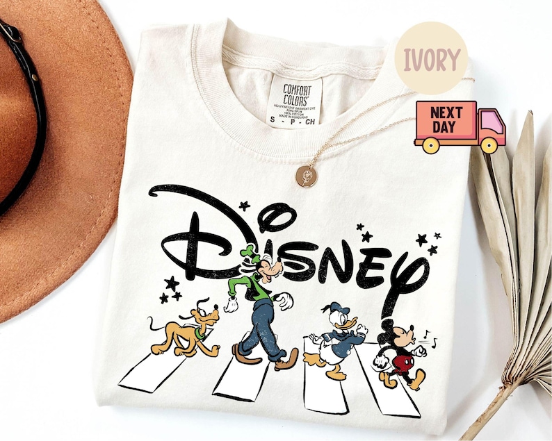 K&ouml;nnte beinhalten: Elfenbeinfarbenes T-Shirt mit dem Wort "Disney" in einer stilisierten Schriftart und Zeichentrickfiguren, die einen Zebrastreifen &uuml;berqueren. Zu den Figuren geh&ouml;ren Goofy, Pluto, Donald Duck und Mickey Mouse. Das Shirt hat einen Rundhalsausschnitt und kurze &Auml;rmel.