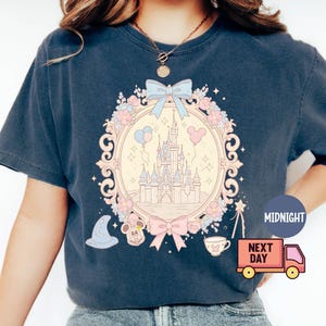K&ouml;nnte beinhalten: Ein marineblaues T-Shirt mit einer pastellrosa und blauen Illustration von Schloss Aschenputtel, umgeben von Blumen und einer Schleife. Der Text "Midnight" und "Next Day" ist auf dem Bild zu sehen.
