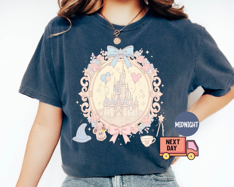 K&ouml;nnte beinhalten: Ein marineblaues T-Shirt mit einer pastellrosa und blauen Illustration von Schloss Aschenputtel, umgeben von Blumen und einer Schleife. Der Text "Midnight" und "Next Day" ist auf dem Bild zu sehen.