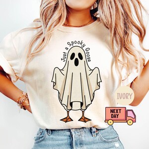 May include: Ivory-colored t-shirt featuring a cartoon ghost with the text "Just a Spooky Goose." The ghost has black eyes and a beak, with brown feet. The shirt is a casual, comfortable style.