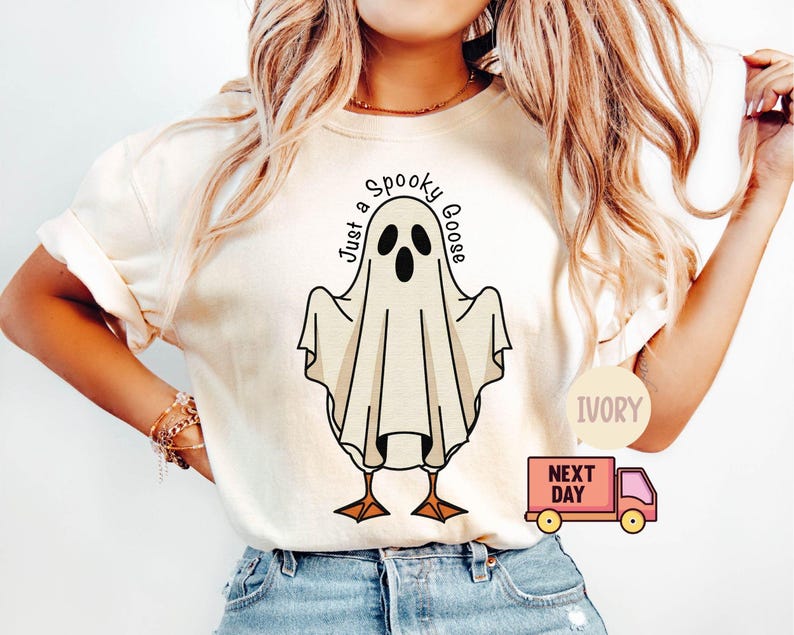 May include: Ivory-colored t-shirt featuring a cartoon ghost with the text "Just a Spooky Goose." The ghost has black eyes and a beak, with brown feet. The shirt is a casual, comfortable style.