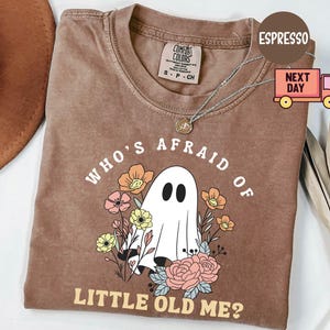 May include: Espresso-colored t-shirt with the phrase "WHO'S AFRAID OF LITTLE OLD ME?" and a graphic of a ghost surrounded by flowers. The shirt has a crew neck and short sleeves. A silver necklace is draped on the shirt.