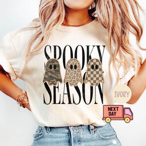 May include: An ivory short-sleeve t-shirt featuring the black text SPOOKY SEASON and three ghost illustrations. The ghosts have distinct patterns: one in camouflage, one in leopard print, and one in a tan and brown checkerboard design.