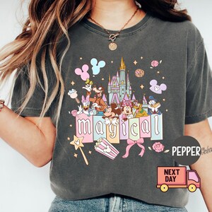 May include: Dark gray t-shirt featuring a colorful graphic with Disney characters and the word "magical." The design includes Mickey and Minnie Mouse, Donald Duck, and a castle. A popcorn box and a magic wand are also included.