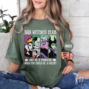 May include: Olive green t-shirt with the text "BAD WITCHES CLUB" and the phrase "WHY BE A PRINCESS WHEN YOU COULD BE A QUEEN?". The shirt features cartoon images of four villainous women in colorful panels. A small pink truck graphic is also visible.