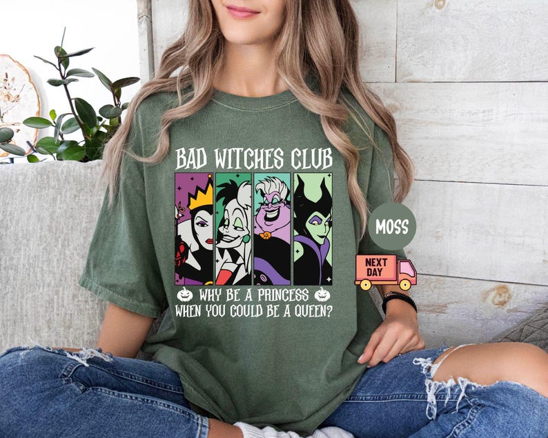 May include: Olive green t-shirt with the text "BAD WITCHES CLUB" and the phrase "WHY BE A PRINCESS WHEN YOU COULD BE A QUEEN?". The shirt features cartoon images of four villainous women in colorful panels. A small pink truck graphic is also visible.