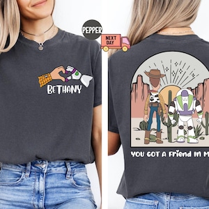 Pu&ograve; includere: Una t-shirt grigio antracite con una grafica frontale di due mani che formano un cuore con il nome BETHANY sotto. La grafica sul retro mostra una scena nel deserto con due personaggi dei cartoni animati e il testo YOU GOT A FRIEND IN ME.