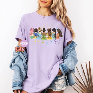 Puede incluir: Camiseta morada con un gr&aacute;fico de siete princesas de pie juntas. Las princesas llevan vestidos de diferentes colores y tienen diferentes peinados. El gr&aacute;fico est&aacute; rodeado de estrellas.