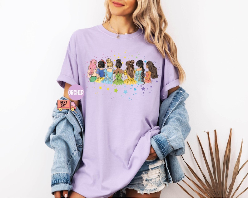 Puede incluir: Camiseta morada con un gr&aacute;fico de siete princesas de pie juntas. Las princesas llevan vestidos de diferentes colores y tienen diferentes peinados. El gr&aacute;fico est&aacute; rodeado de estrellas.