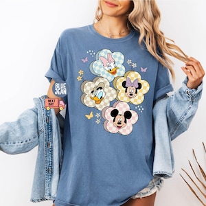 May include: Blue crew neck t-shirt featuring a floral design with Disney characters. The flower petals have a checkered pattern and feature Donald Duck, Daisy Duck, Minnie Mouse, and Mickey Mouse. The shirt has rolled-up sleeves and the text "BLUE JEAN" is visible.