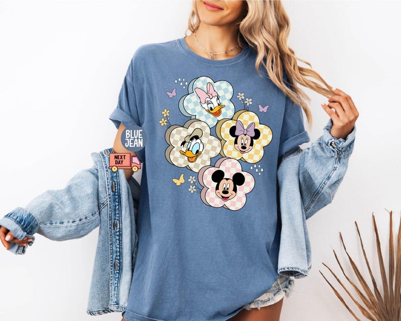 May include: Blue crew neck t-shirt featuring a floral design with Disney characters. The flower petals have a checkered pattern and feature Donald Duck, Daisy Duck, Minnie Mouse, and Mickey Mouse. The shirt has rolled-up sleeves and the text "BLUE JEAN" is visible.