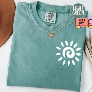 May include: Light green Comfort Colors t-shirt with a white sun graphic. The shirt has a crew neck and short sleeves. A gold necklace with a pendant is on the shirt. The shirt is labeled "Light Green" and "Next Day".
