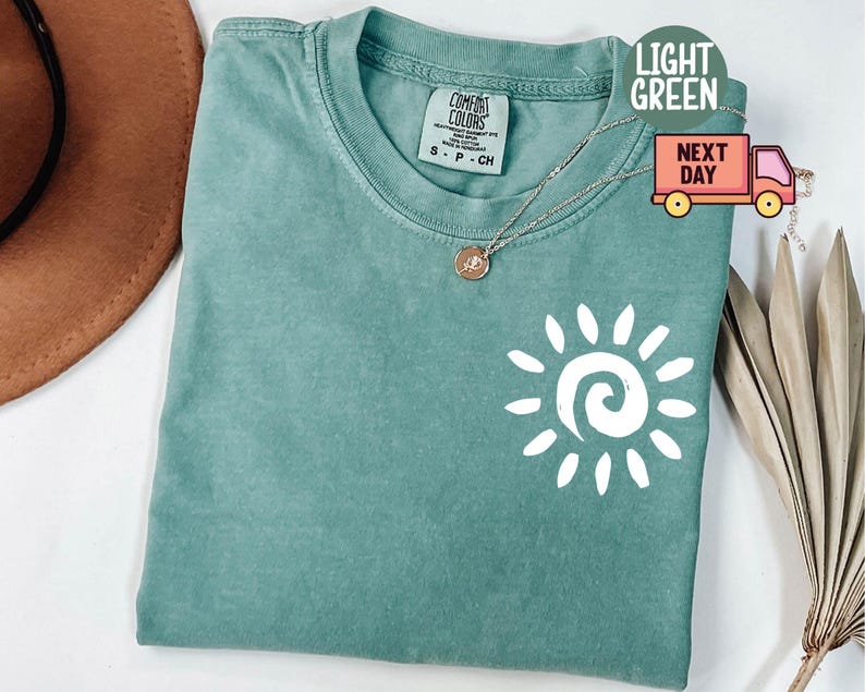 May include: Light green Comfort Colors t-shirt with a white sun graphic. The shirt has a crew neck and short sleeves. A gold necklace with a pendant is on the shirt. The shirt is labeled "Light Green" and "Next Day".