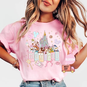 May include: Pink t-shirt featuring a colorful graphic of Disney characters, a castle, and the word "magical". The design includes Mickey Mouse, Minnie Mouse, Donald Duck, and other characters. A pink bow and a magic wand are also included in the design.