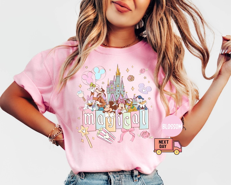May include: Pink t-shirt featuring a colorful graphic of Disney characters, a castle, and the word "magical". The design includes Mickey Mouse, Minnie Mouse, Donald Duck, and other characters. A pink bow and a magic wand are also included in the design.