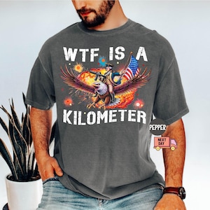 May include: Dark gray t-shirt with a graphic featuring an eagle carrying a person holding a rifle and an American flag. The text "WTF IS A KILOMETER" is printed in white.