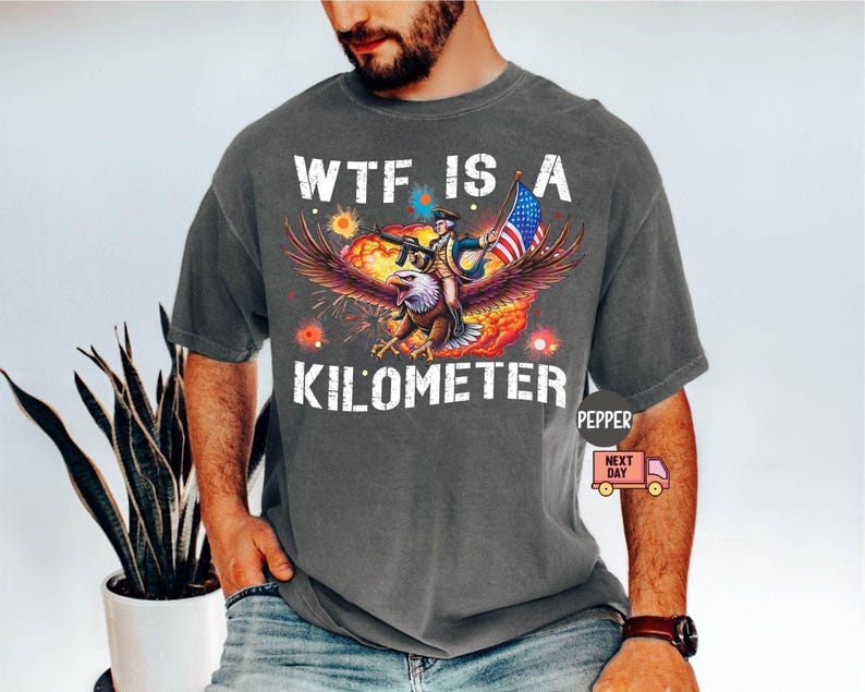 May include: Dark gray t-shirt with a graphic featuring an eagle carrying a person holding a rifle and an American flag. The text "WTF IS A KILOMETER" is printed in white.