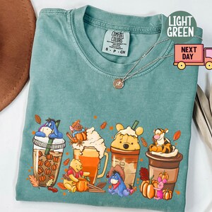 May include: Light green Comfort Colors t-shirt featuring a graphic of Winnie the Pooh characters as fall-themed drinks. The design includes Eeyore, Piglet, Tigger, and Pooh Bear in coffee cups with pumpkins and fall leaves.