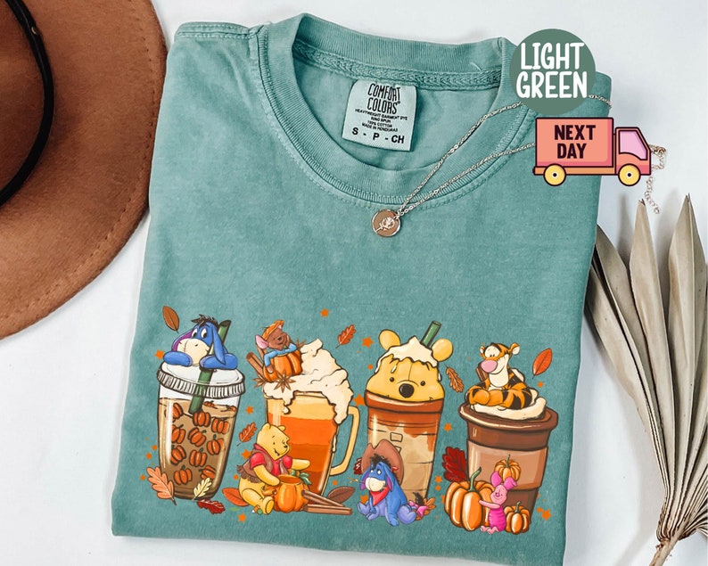 May include: Light green Comfort Colors t-shirt featuring a graphic of Winnie the Pooh characters as fall-themed drinks. The design includes Eeyore, Piglet, Tigger, and Pooh Bear in coffee cups with pumpkins and fall leaves.