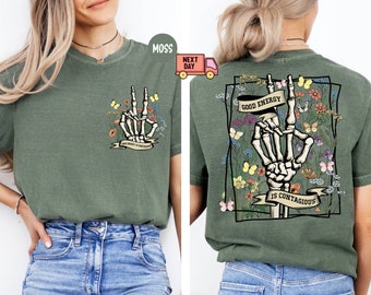 May include: Green t-shirt with a skeleton hand making a peace sign surrounded by flowers and butterflies. The text on the shirt reads "Good Energy is Contagious".