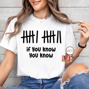 May include: White t-shirt with black tally marks and the phrase "if you know you know." The shirt has a crew neck and short sleeves. The text is in a bold, sans-serif font.