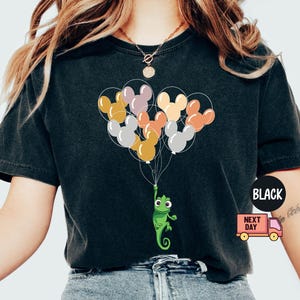 Może przedstawiać: Czarny T-shirt z zielonym kameleonem trzymającym wiązkę kolorowych balon&oacute;w z uszami Myszki Miki. Balony są ż&oacute;łte, pomarańczowe, czerwone, r&oacute;żowe i fioletowe.